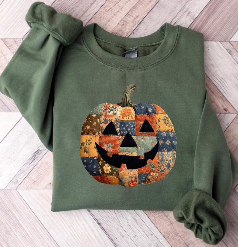 May include: Olive green sweatshirt featuring a patchwork pumpkin design. The pumpkin has a smiling face and is made of various floral and patterned fabrics in shades of orange, blue, and yellow. The sleeves are cuffed.