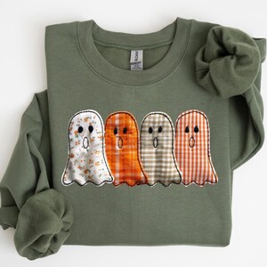 May include: An olive green sweatshirt featuring four ghost appliqués. Each ghost has a different patterned fabric: floral, orange plaid, tan plaid, and orange and white plaid. The ghosts have black dot eyes and a stitched mouth.