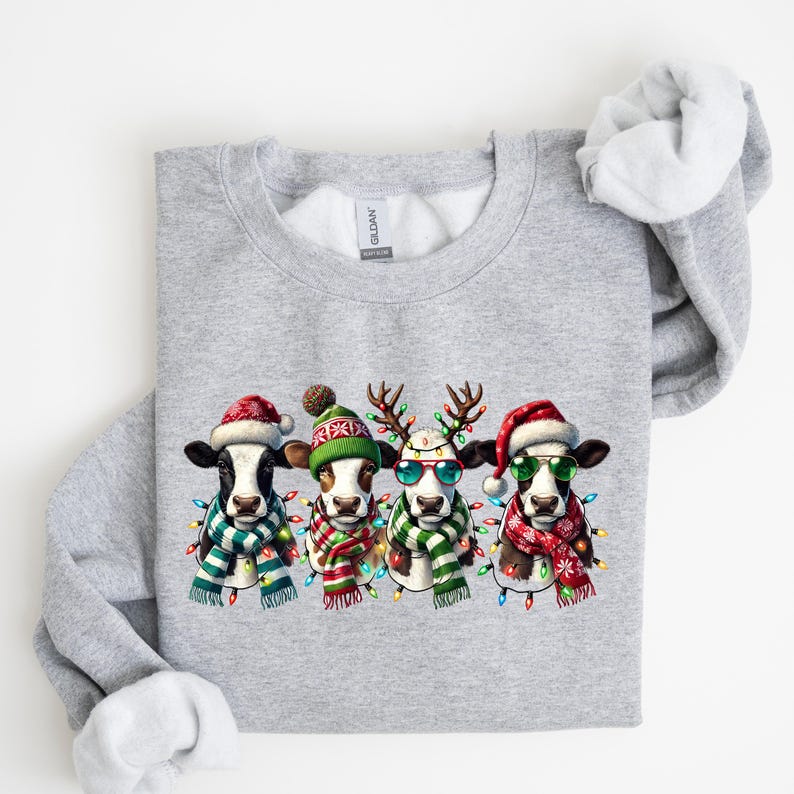 K&ouml;nnte beinhalten: Hellgraues Sweatshirt mit festlichem Design von vier K&uuml;hen. Jede Kuh tr&auml;gt einen Weihnachtsmannhut, einen Schal und eine Lichterkette, eine Kuh tr&auml;gt Rentiergeweih und eine Sonnenbrille. Die K&uuml;he sind in einer Reihe angeordnet.
