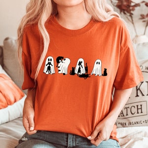 May include: A bright orange t-shirt featuring a graphic of five ghosts and black cats. The ghosts are white with black eyes and mouths. The cats are black with white eyes. The graphic is centred on the shirt.