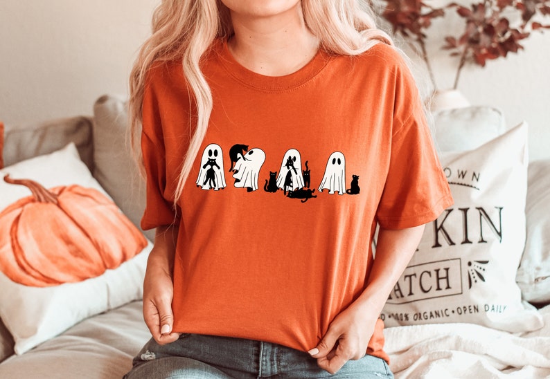 May include: A bright orange t-shirt featuring a graphic of five ghosts and black cats. The ghosts are white with black eyes and mouths. The cats are black with white eyes. The graphic is centred on the shirt.