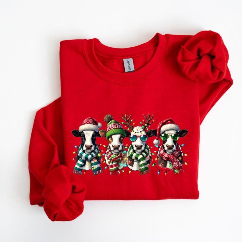 K&ouml;nnte beinhalten: Ein rotes Sweatshirt mit vier Cartoon-K&uuml;hen, die f&uuml;r Weihnachten gekleidet sind. Jede Kuh tr&auml;gt einen festlichen Hut, einen Schal und eine Sonnenbrille mit Lichterketten. Die K&uuml;he sind schwarz-wei&szlig; mit braunen Flecken.