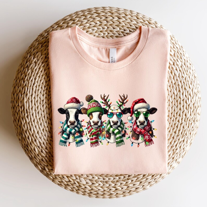 K&ouml;nnte beinhalten: Hellrosa T-Shirt mit vier Cartoon-K&uuml;hen, die f&uuml;r Weihnachten gekleidet sind. Jede Kuh tr&auml;gt einen Weihnachtsmannhut, einen Schal und eine Lichterkette. Eine Kuh hat Rentiergeweih und eine Sonnenbrille. Das T-Shirt ist auf einer gewebten Oberfl&auml;che gefaltet.