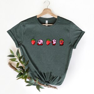 Pu&ograve; includere: T-shirt verde scuro con un disegno di personaggi dei cartoni animati a forma di fragole. I personaggi hanno corpi rosa, cime di fragole rosse e occhi neri. La maglietta &egrave; appesa a una gruccia di legno.