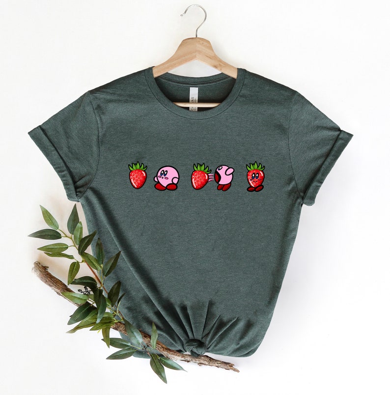 Pu&ograve; includere: T-shirt verde scuro con un disegno di personaggi dei cartoni animati a forma di fragole. I personaggi hanno corpi rosa, cime di fragole rosse e occhi neri. La maglietta &egrave; appesa a una gruccia di legno.