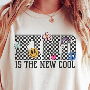 May include: A white t-shirt with a black and white checkered design that spells out "KIND IS THE NEW COOL". The letters are decorated with colorful flowers, a smiley face, a rainbow, a peace sign, and a heart.
