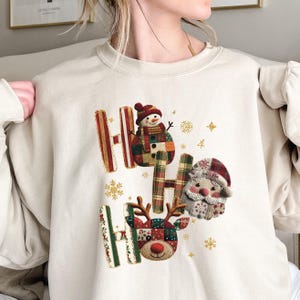 May include: Cream-coloured sweatshirt with a festive "HO HO HO" design. The design features a snowman, Santa Claus, and a reindeer, all in a patchwork style. Gold snowflakes and accents complete the holiday theme.
