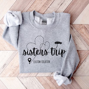 May include: Gray sweatshirt with the text "sisters-trip" in black lettering. A car icon and a pin icon are above the text, with a dotted line connecting them. The text "CUSTOM LOCATION" is below the text "sisters-trip".