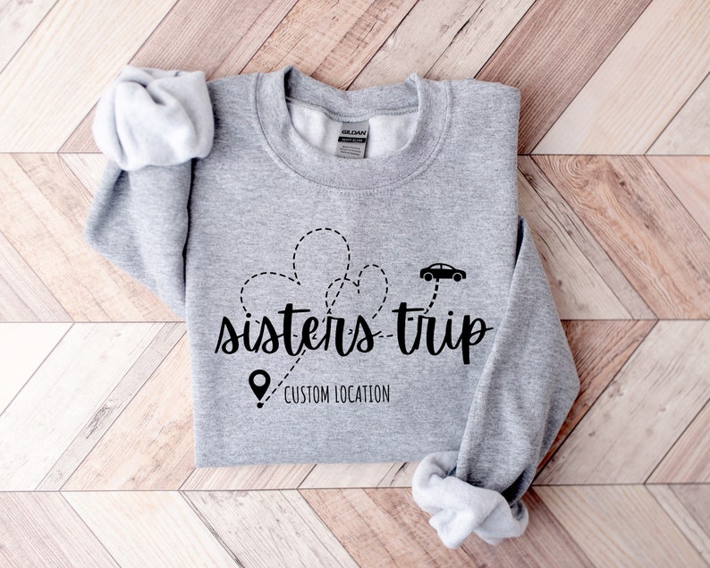 May include: Gray sweatshirt with the text "sisters-trip" in black lettering. A car icon and a pin icon are above the text, with a dotted line connecting them. The text "CUSTOM LOCATION" is below the text "sisters-trip".