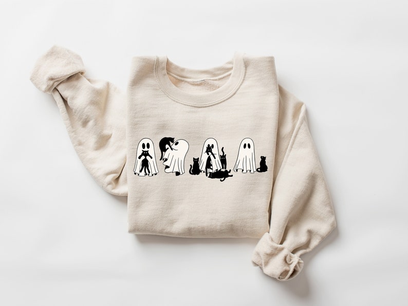 May include: A beige sweatshirt featuring a black and white graphic of cats dressed as ghosts. The design shows five cats, each with a ghost sheet over their head. The cats are in various poses, including sitting, standing, and playing.