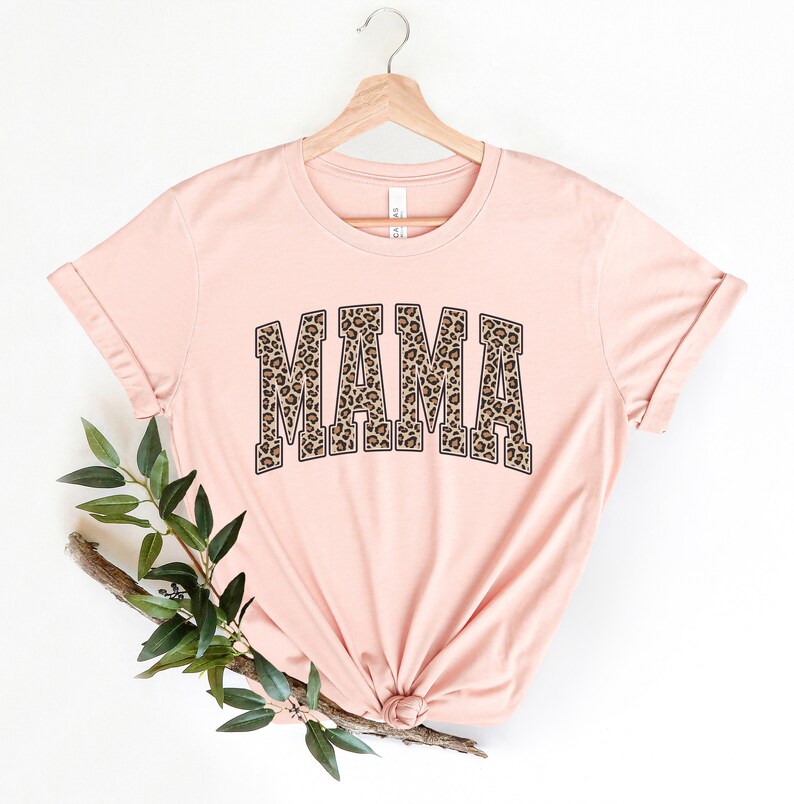 May include: A pink t-shirt with a leopard print "MAMA" graphic. The shirt is on a hanger and has a relaxed fit.