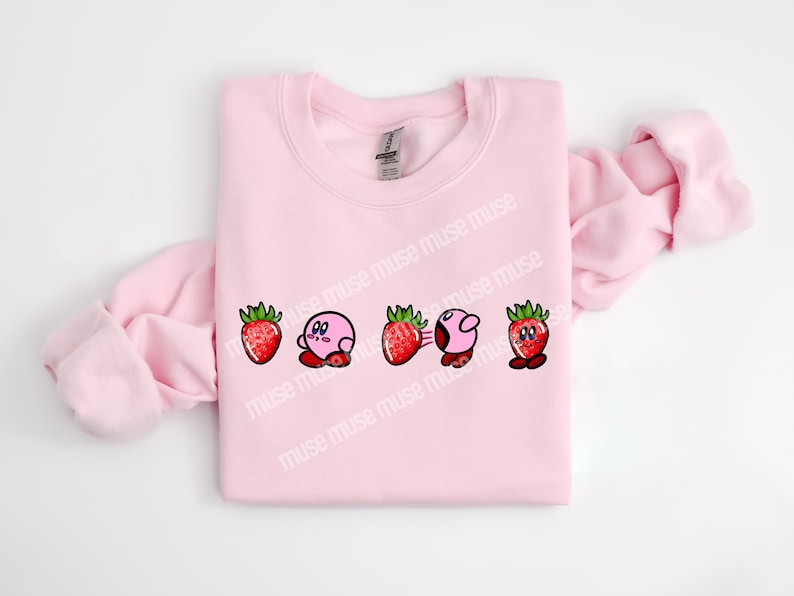 Pu&ograve; includere: Una felpa rosa chiaro con un design giocoso di un personaggio dei cartoni animati e fragole. Il design comprende cinque immagini: una fragola, un personaggio rosa, una fragola, il personaggio rosa con linee di movimento e una fragola.