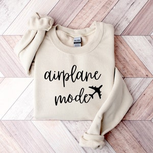 Puede incluir: Una sudadera beige con el texto "airplane mode" en letras cursivas negras y un peque&ntilde;o gr&aacute;fico de avi&oacute;n.