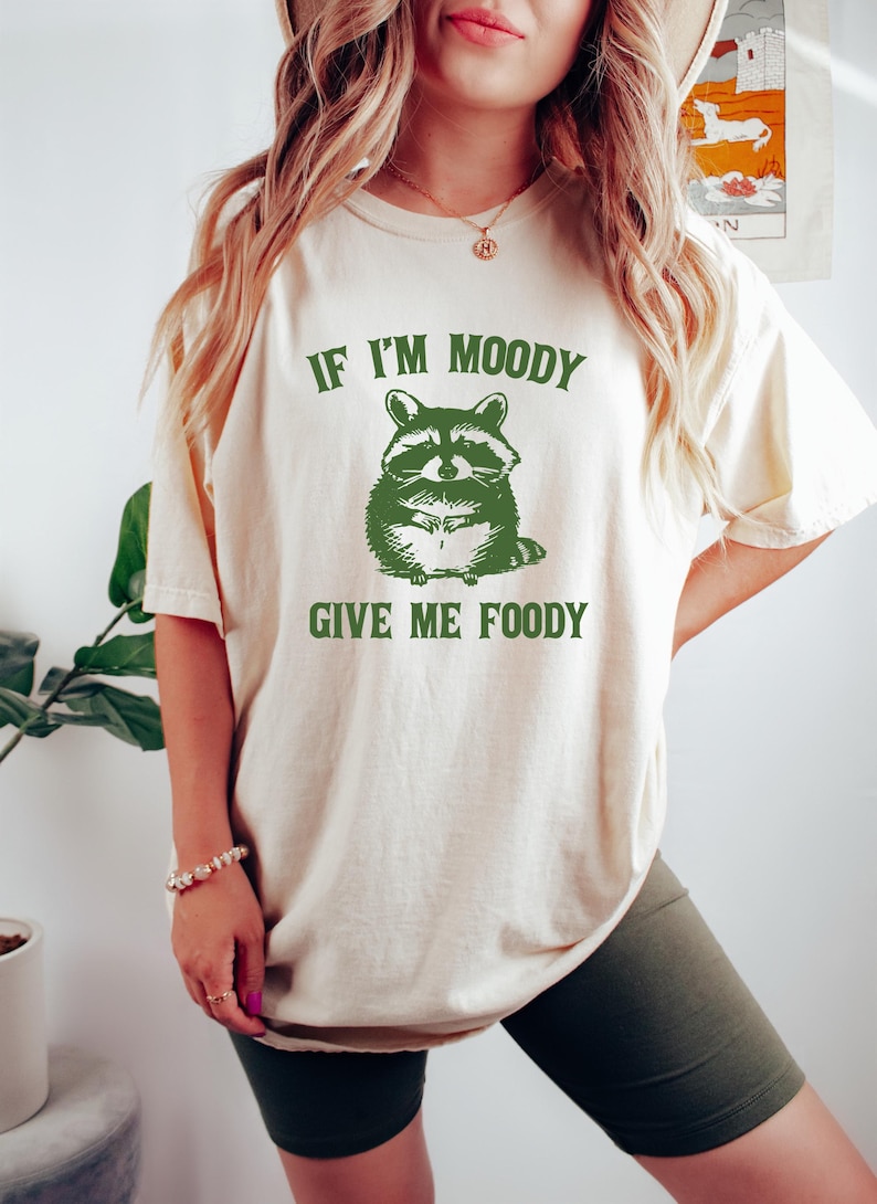 May include: A white t-shirt with a green raccoon graphic and the text "If I'm Moody Give Me Foody".