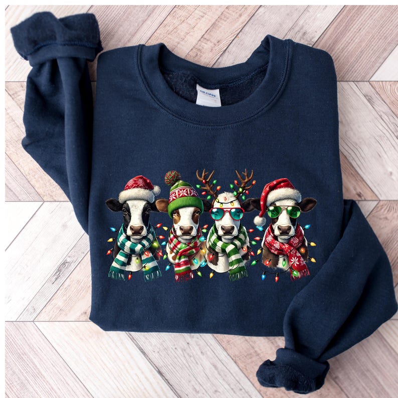 K&ouml;nnte beinhalten: Marineblaues Sweatshirt mit einem festlichen Design. Es zeigt vier K&uuml;he, die f&uuml;r die Feiertage gekleidet sind. Jede Kuh tr&auml;gt einen Weihnachtsm&uuml;tze, einen Schal und eine Lichterkette. Eine Kuh tr&auml;gt eine Sonnenbrille und eine andere ein Rentiergeweih.