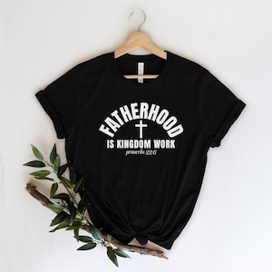 May include: Black t-shirt with the white text "FATHERHOOD is KINGDOM WORK" and a cross graphic. The text "proverbs 22:6" is below the main text. The shirt is displayed on a wooden hanger.