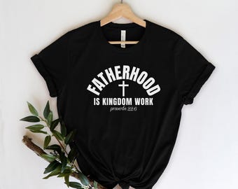 May include: Black t-shirt with the white text "FATHERHOOD is KINGDOM WORK" and a cross graphic. The text "proverbs 22:6" is below the main text. The shirt is displayed on a wooden hanger.