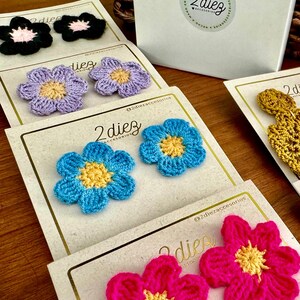 May include: Assortment of crocheted flower embellishments in various colors, including pink, blue, yellow, and purple. The flowers are displayed on white cards with the text "2diez accesorios" and "@2diezaccesorios".