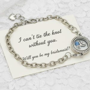 May include: Silver-toned charm bracelet with a round locket containing a blue gem and a heart charm. The bracelet is on a card that reads "I can't tie the knot without you. Will you be my bridesmaid?"