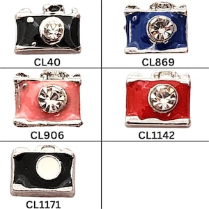 May include: Five miniature camera charms in black, blue, pink, and red, each with a sparkling crystal lens. The charms have silver-colored metal detailing and are labeled with the codes CL40, CL869, CL906, CL1142, and CL1171.