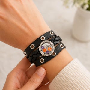 May include: Black leather wrap bracelet with silver-tone grommets and a braided section. The bracelet features a round charm with orange, purple, and clear accents. A stylish accessory for everyday wear.