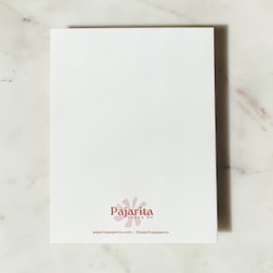 May include: A white rectangular card with the Pajartia Paper Co. logo in the bottom center. The logo is a stylized flower in a light pink color. The card is on a white and gray marble surface. The website and social media handle are printed below the logo.