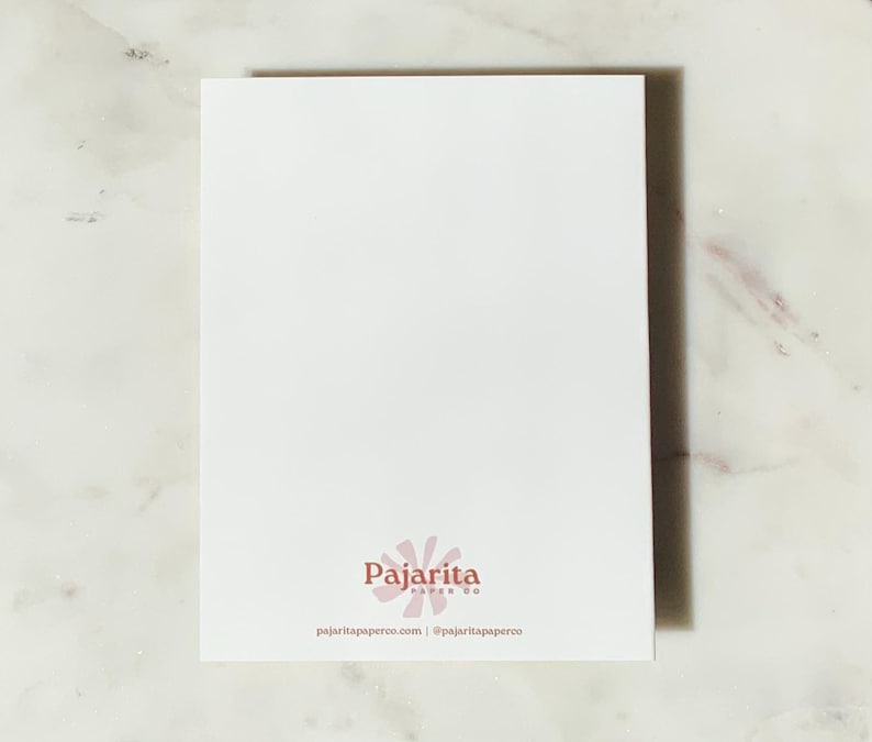 May include: A white rectangular card with the Pajartia Paper Co. logo in the bottom center. The logo is a stylized flower in a light pink color. The card is on a white and gray marble surface. The website and social media handle are printed below the logo.