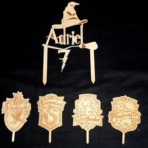 May include: Wooden cake toppers featuring the Sorting Hat, a wand, the name "Adriel", the number "7", and the four Hogwarts house crests: Gryffindor, Hufflepuff, Slytherin, and Ravenclaw.
