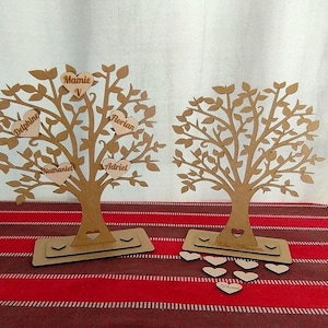 Peut inclure: Deux décorations d'arbre généalogique en bois avec des branches et des cœurs découpés. Les cœurs portent des noms écrits dessus, dont "Mamie V", "Delphine", "Nathaniel", "Florian" et "Adriel". Un arbre a le texte "Notre Famille" sur la base.