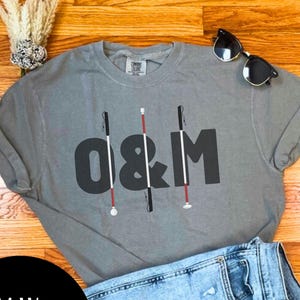 May include: A grey short-sleeve t-shirt featuring the black text O&M with three vertical mobility canes integrated into the letters. The shirt is paired with light blue acid-wash denim shorts and black sunglasses resting on the fabric.