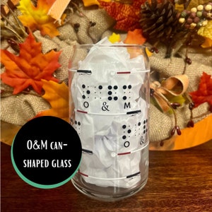 May include: A clear glass can-shaped drinking glass with a white background and black braille dots that spell out "O & M". The glass is sitting on a wooden surface with fall foliage in the background.