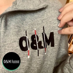 O&M white cane fleece! The design has large, bold O&M print with three white cane overlays. The embroidery is beautiful and stands out perfectly against the grey fleece.