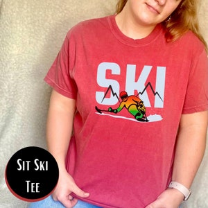 May include: Red t-shirt with a white graphic of a skier and the word "SKI" with mountains. The text "SIT SKI TEE" is in a black circle.