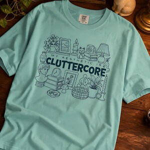 May include: A light mint green short-sleeve t-shirt featuring a dark blue graphic print of various household objects and the text "MY AESTHETIC IS CLUTTERCORE". The design includes illustrations of a bust, books, a clock, plants, and a cat.