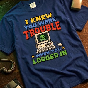Può includere: Una t-shirt blu navy con una grafica in pixel art di un computer vintage con un teschio sullo schermo. Il testo recita I KNEW YOU WERE TROUBLE WHEN YOU LOGGED IN, con lettere pixelate di colore giallo, rosso e verde.