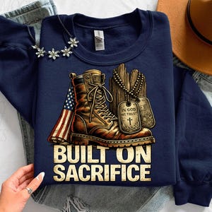 May include: Navy blue sweatshirt with a graphic featuring a brown combat boot, an American flag, and dog tags. The text "BUILT ON SACRIFICE" is displayed in a bold, gold font. A necklace with flower-shaped pendants is visible.