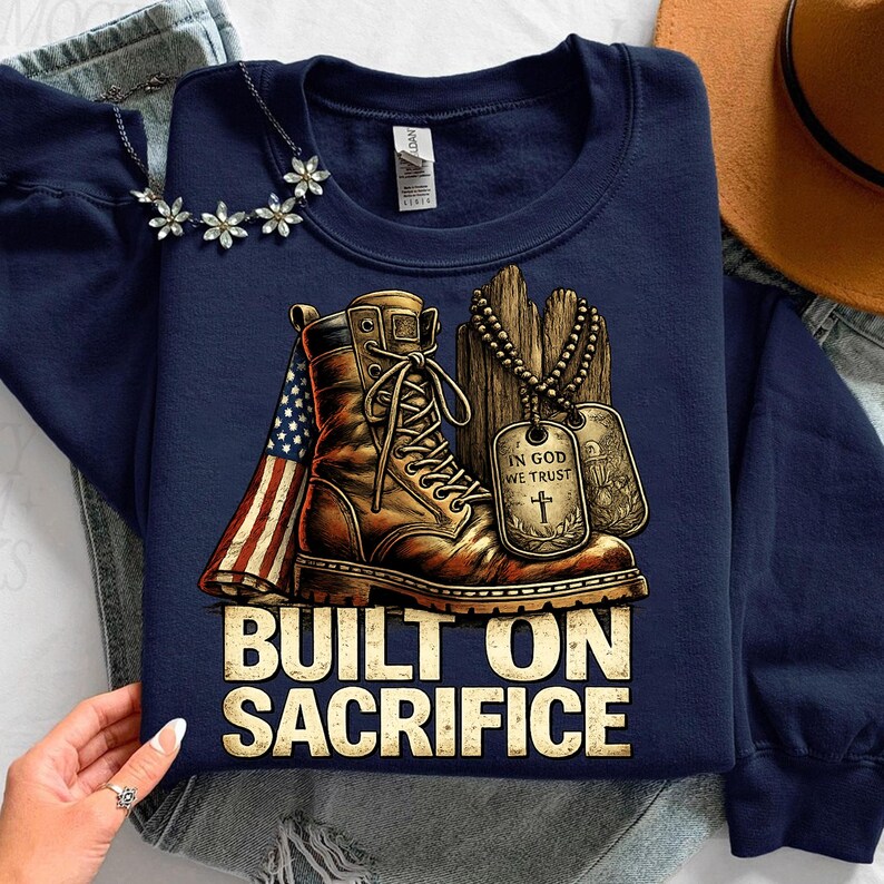 May include: Navy blue sweatshirt with a graphic featuring a brown combat boot, an American flag, and dog tags. The text "BUILT ON SACRIFICE" is displayed in a bold, gold font. A necklace with flower-shaped pendants is visible.