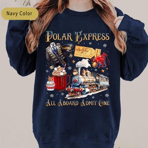 May include: Navy blue sweatshirt with a colorful graphic of a train, a cup of hot chocolate, and a ticket. The text "Polar Express" and "All Aboard Admit One" are also displayed. The design includes snowflakes and stars.
