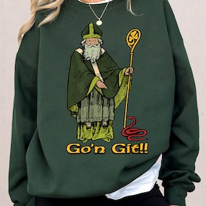 May include: Dark green sweatshirt featuring a vintage illustration of a saintly figure in green robes, holding a staff with a shamrock. A red snake is at the bottom. The text "Go'n Git!!" is in yellow. The sweatshirt is a casual garment.