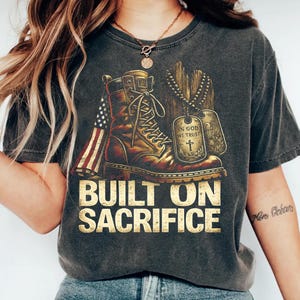 May include: Dark gray t-shirt featuring a distressed graphic of a combat boot, American flag, and dog tags. The text "BUILT ON SACRIFICE" is printed in gold. The shirt has a vintage, worn look.
