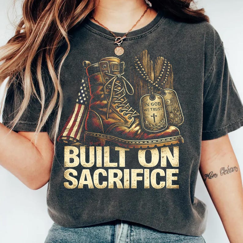 May include: Dark gray t-shirt featuring a distressed graphic of a combat boot, American flag, and dog tags. The text "BUILT ON SACRIFICE" is printed in gold. The shirt has a vintage, worn look.