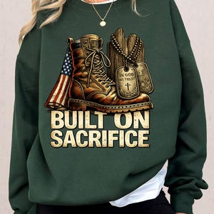 May include: Dark green sweatshirt featuring a graphic of a combat boot, dog tags, and an American flag. The text "BUILT ON SACRIFICE" is displayed in a bold, gold font. The design evokes themes of military service and patriotism.