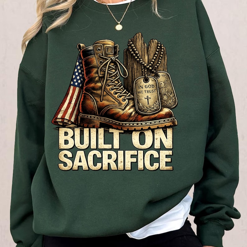 May include: Dark green sweatshirt featuring a graphic of a combat boot, dog tags, and an American flag. The text "BUILT ON SACRIFICE" is displayed in a bold, gold font. The design evokes themes of military service and patriotism.