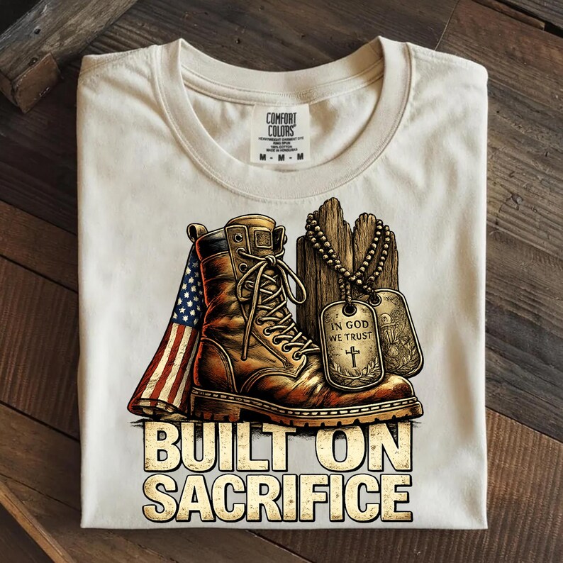 May include: Cream-colored t-shirt featuring a graphic of a combat boot, American flag, and dog tags. The boot is brown with detailed laces. The dog tags read "IN GOD WE TRUST." The text "BUILT ON SACRIFICE" is below the graphic.