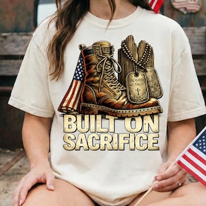 May include: Cream-colored t-shirt featuring a graphic of a brown combat boot, an American flag, and dog tags. The text "BUILT ON SACRIFICE" is displayed below the boot. The design has a vintage, patriotic theme.