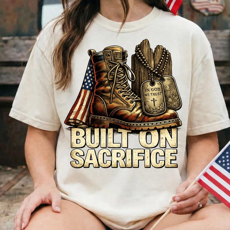 May include: Cream-colored t-shirt featuring a graphic of a brown combat boot, an American flag, and dog tags. The text "BUILT ON SACRIFICE" is displayed below the boot. The design has a vintage, patriotic theme.