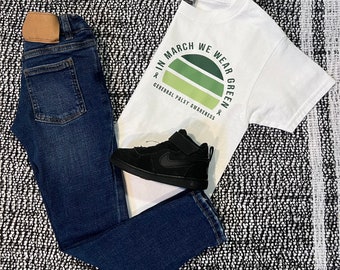 May include: A white t-shirt with the text "In March We Wear Green" and a green circle graphic. The text "General Palsy Awareness" is below the circle. A pair of blue jeans and black sneakers are also pictured.