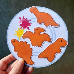 May include: A round sticker featuring four orange dinosaur-shaped nuggets on a light blue plate. Splashes of pink and yellow are also on the plate. The sticker is held by a hand.