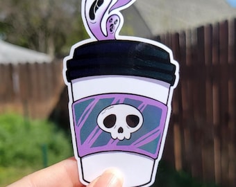 Op de afbeelding: Een witte koffiebeker sticker met een zwart deksel en een paarse band met een schedel ontwerp. Boven de beker zweven paarse spookachtige figuren omhoog. De sticker wordt door een hand vastgehouden.