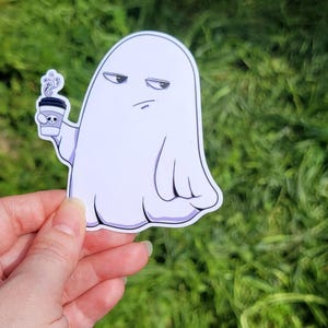 May include: A white sticker featuring a cartoon ghost with a grumpy expression, holding a coffee cup. The cup has a skull design and steam rising from it. The ghost is outlined in black.